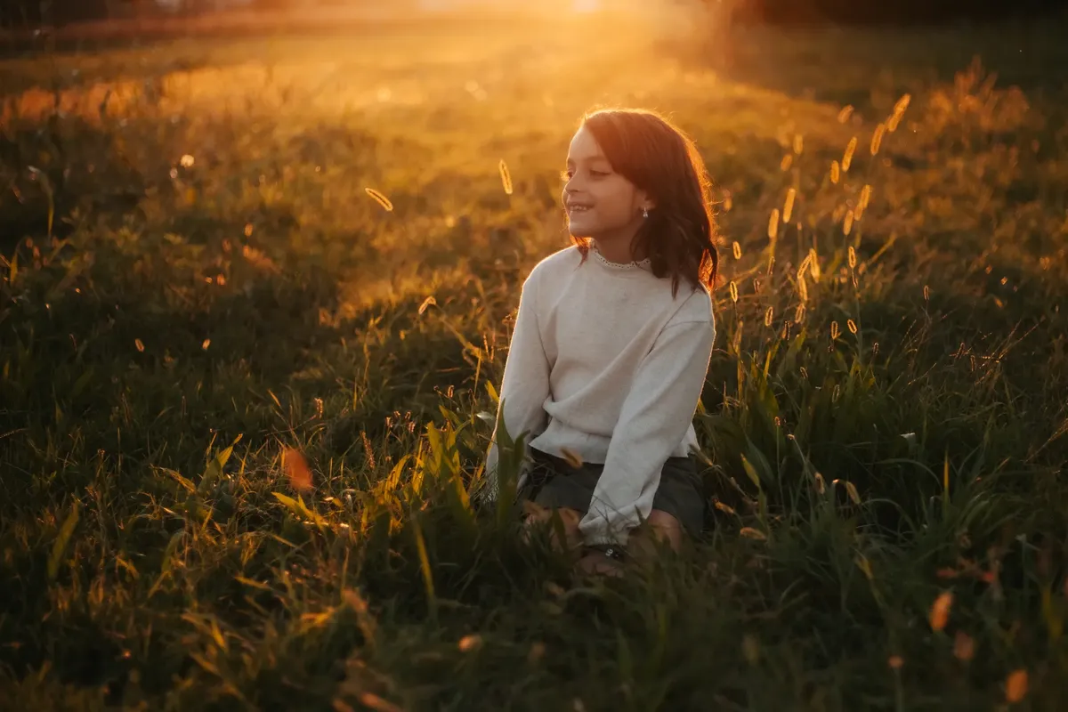 Bambina sorridente seduta nell'erba alta illuminata dal sole al tramonto