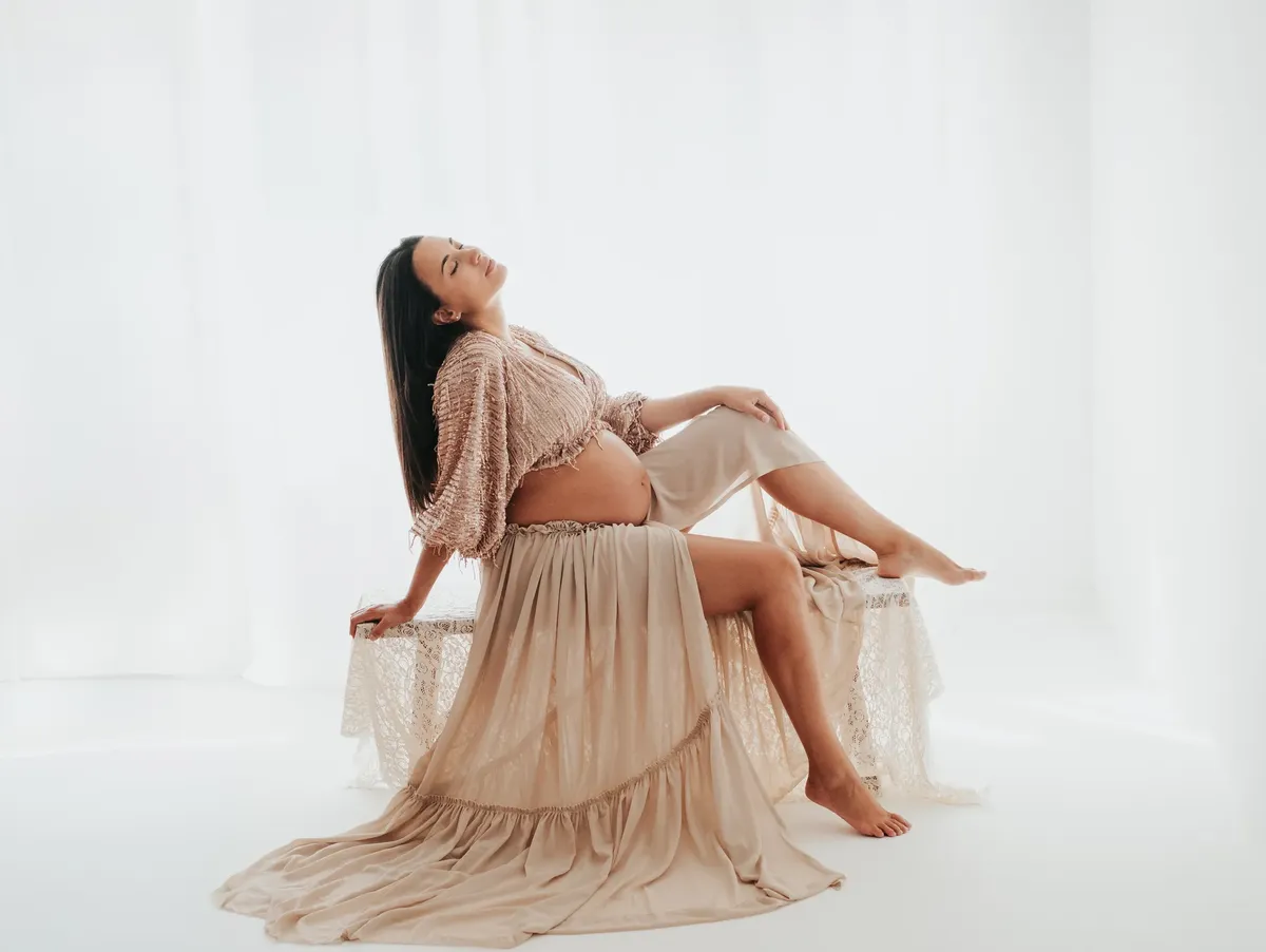 Futura mamma seduta in abito bohémien color cipria con pancione scoperto, servizio maternità in studio