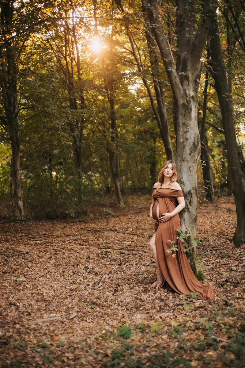 Donna incinta in abito marrone appoggiata a un albero nel bosco autunnale
