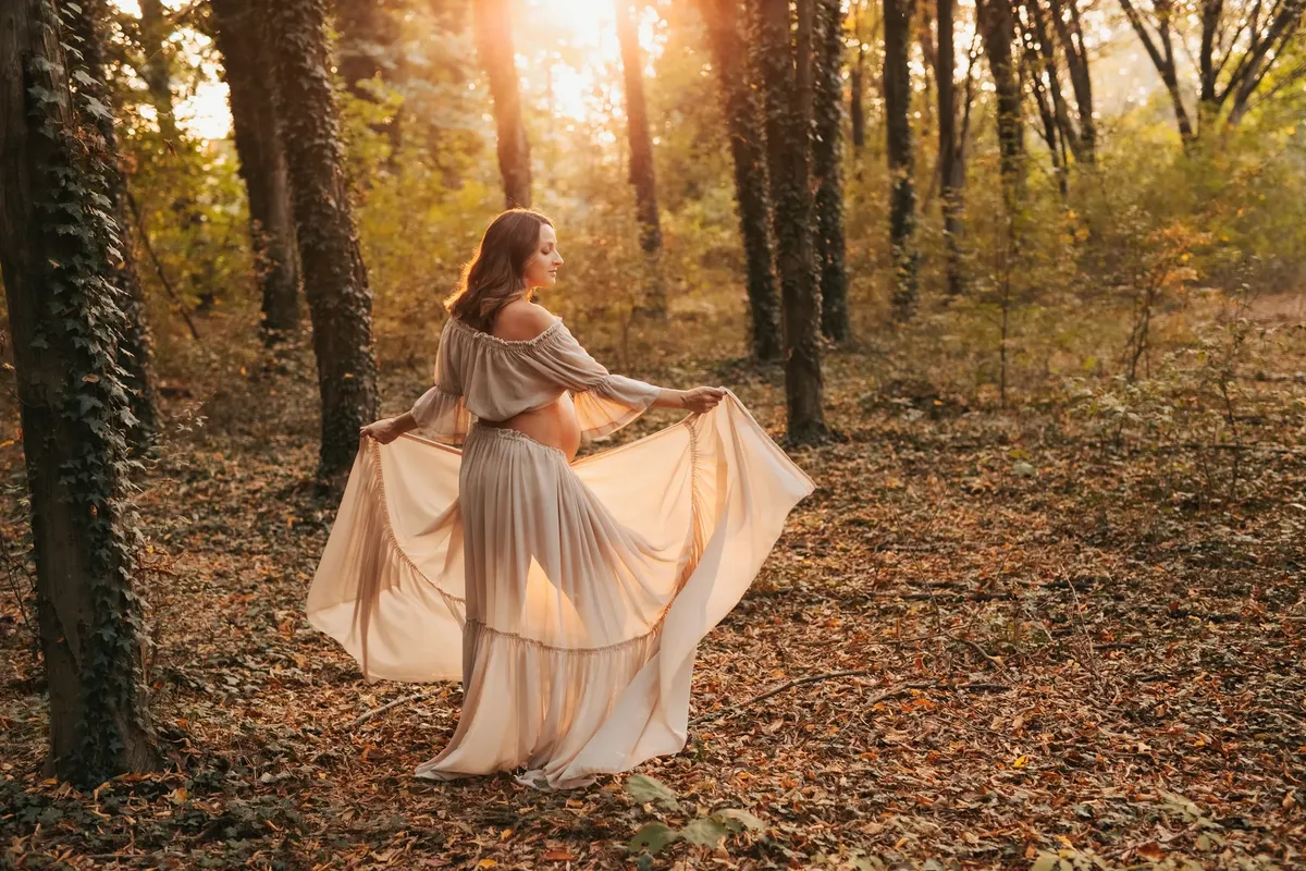 Donna incinta in abito fluido danza nel bosco autunnale con luce dorata tra gli alberi