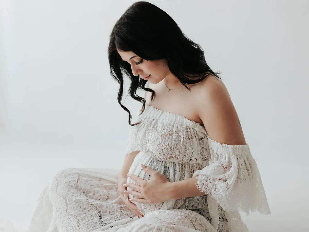 Futura mamma seduta in abito pizzo bianco, guarda dolcemente il pancione in studio