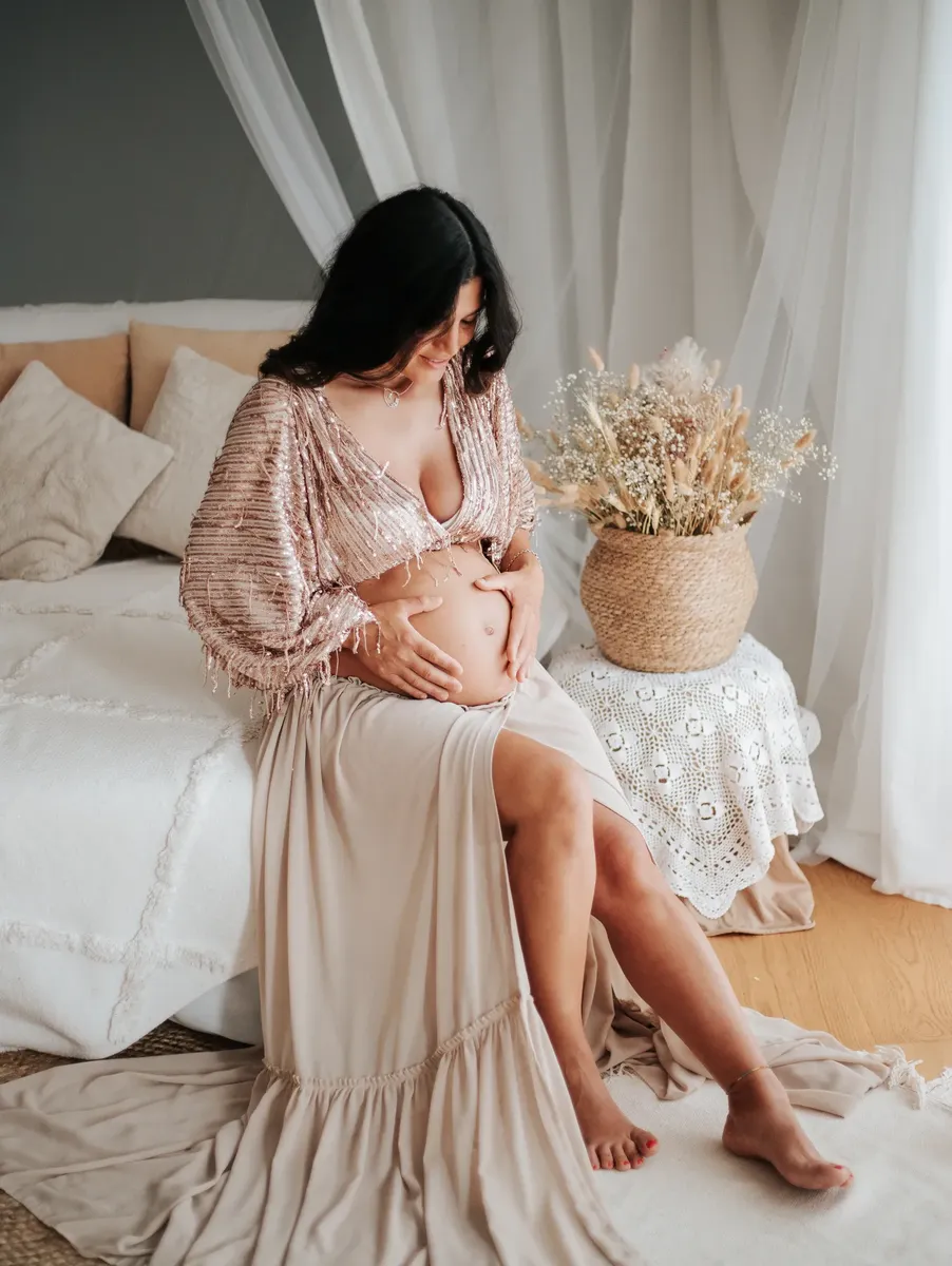 Donna incinta seduta sul letto con top dorato e gonna beige, set boho elegante