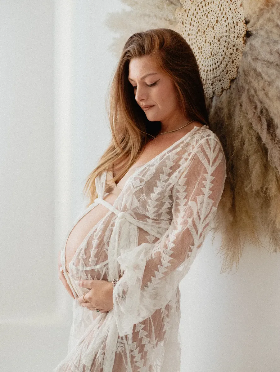 Futura mamma in pizzo bianco tra decorazioni di pampa accarezza il pancione in studio