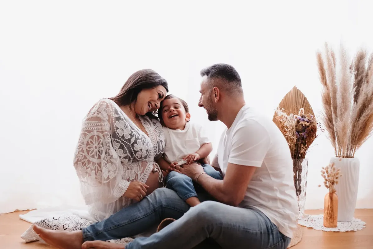 Famiglia premaman con bimbo che ride seduti a terra con fiori secchi e pampa