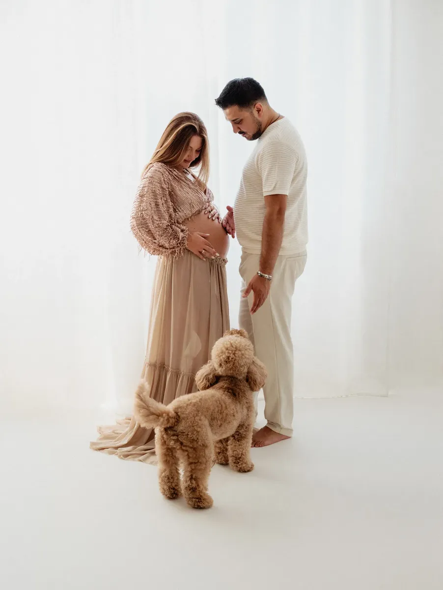 Coppia premaman in studio con cagnolino, lei in abito lungo beige con frange