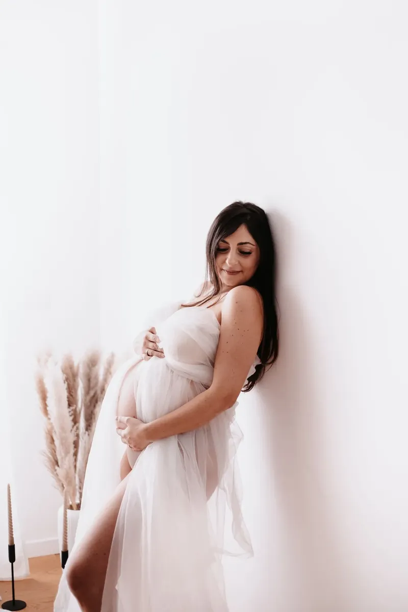 Gestante in abito di tulle bianco appoggiata al muro con pampa decorativa