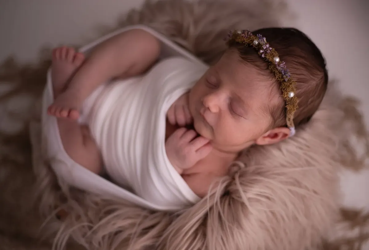 Neonata avvolta in wrap bianco con coroncina floreale su pelliccia beige