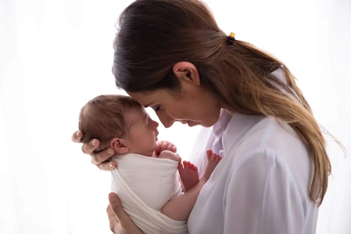 Mamma con neonato avvolto in wrap bianco, naso contro naso su sfondo chiaro