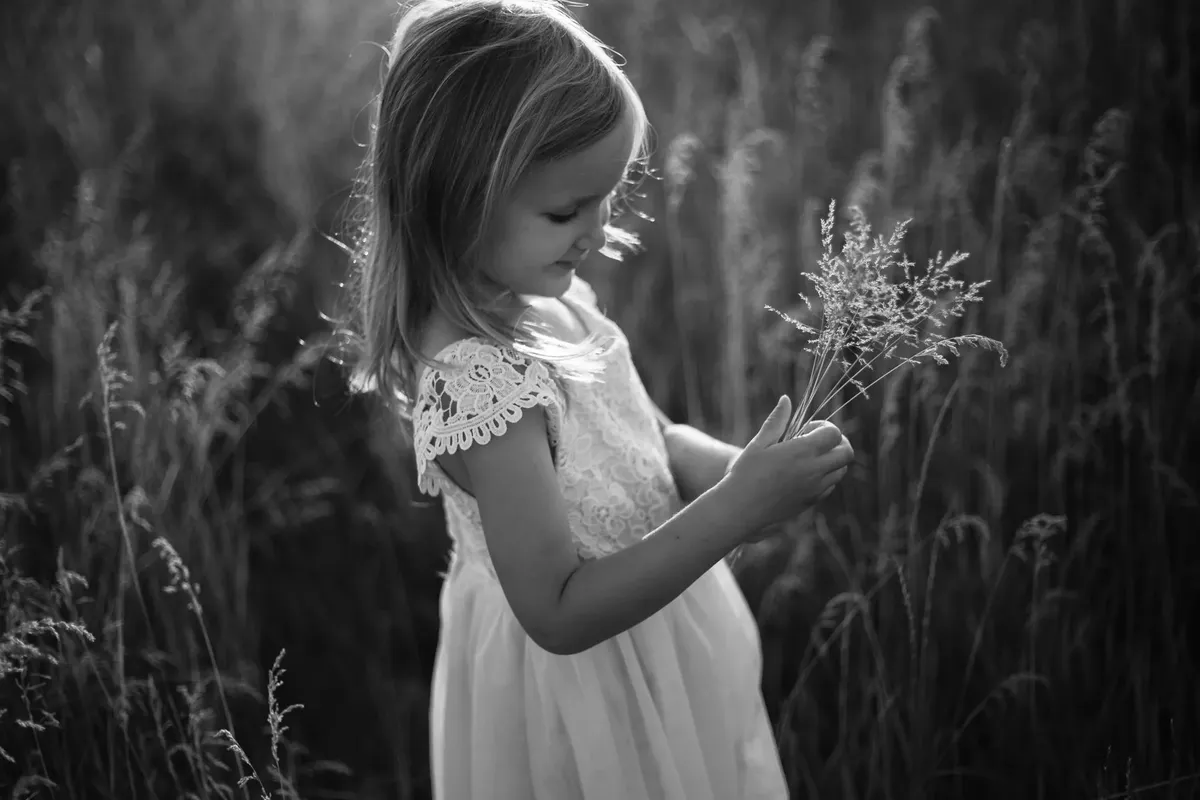 Bambina in abito bianco di pizzo che tiene un mazzolino di fiori di campo, ritratto in bianco e nero controluce