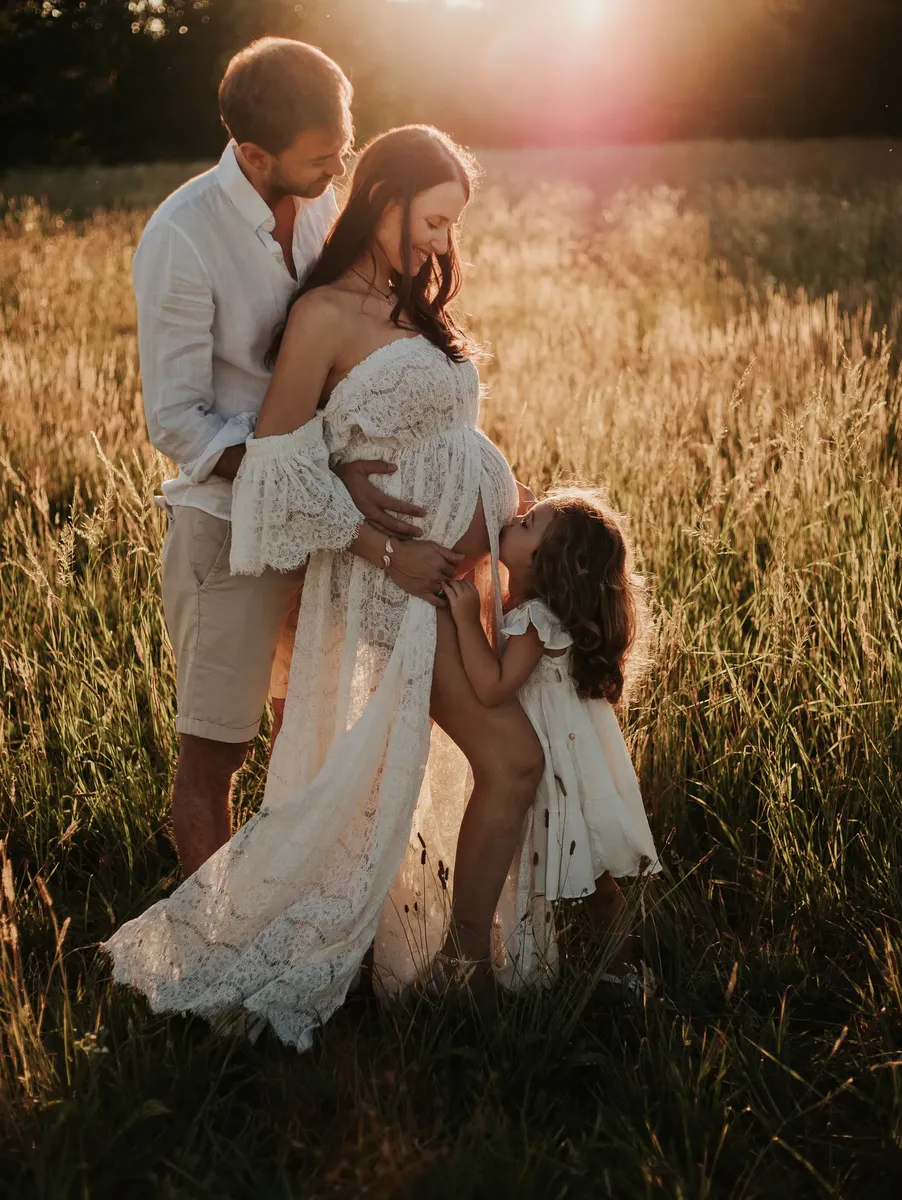 Famiglia al tramonto in un campo, papà abbraccia mamma incinta mentre la figlia bacia il pancione, controluce dorato