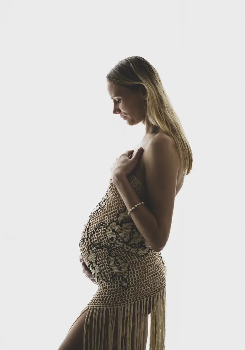 Silhouette di futura mamma di profilo in abito a rete con frange, ritratto artistico controluce in studio