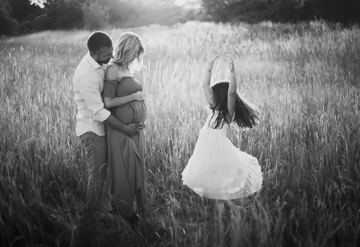 Coppia in attesa abbracciata in un prato al tramonto con bambina che danza, ritratto di famiglia in bianco e nero