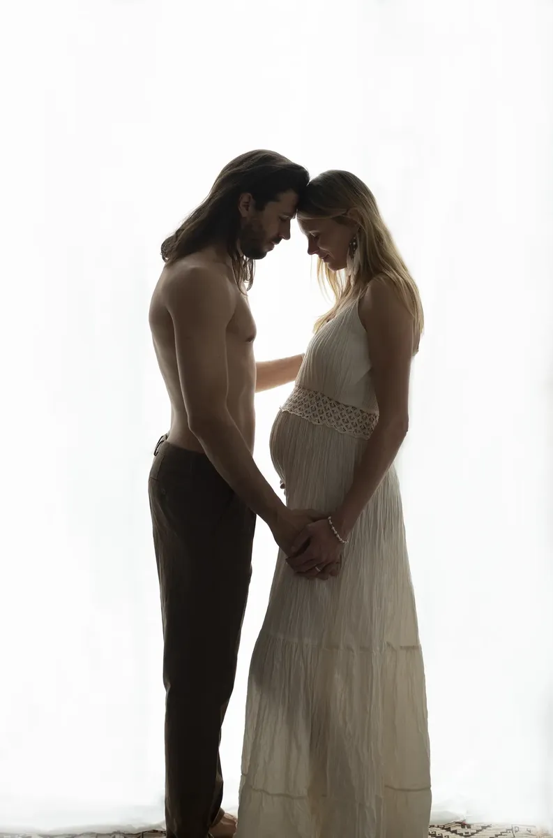 Coppia in attesa fronte contro fronte in controluce, lei in abito lungo chiaro e lui a torso nudo, silhouette romantica
