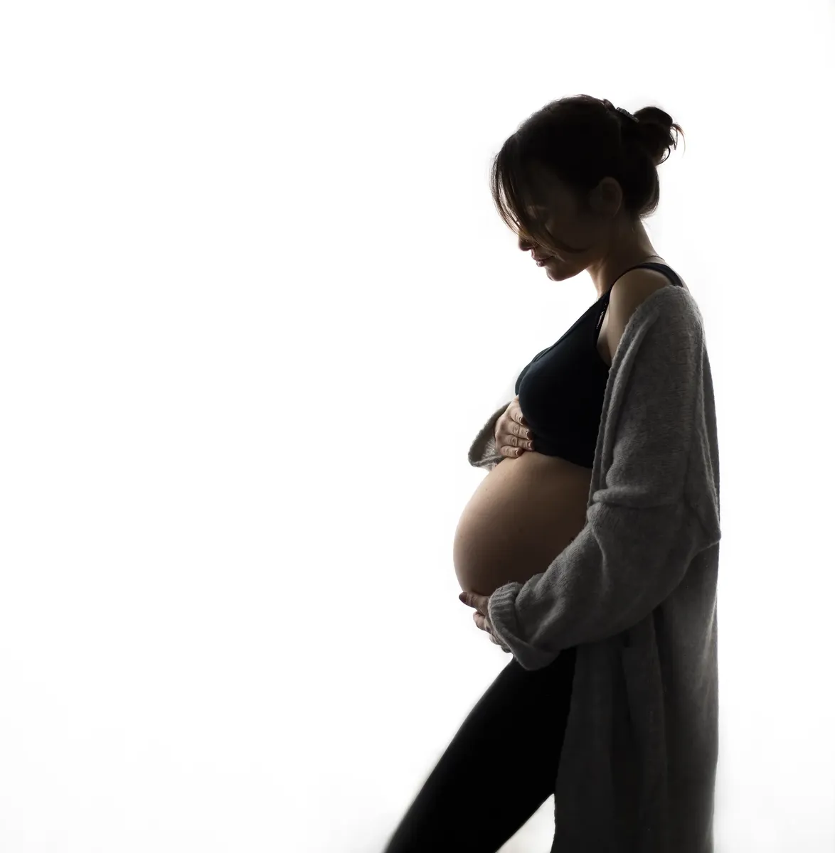 Silhouette di futura mamma di profilo in cardigan grigio che accarezza il pancione, controluce in studio